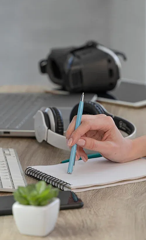 A person's hand writes in a notebook on a desk with a laptop.